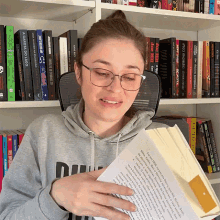 a woman wearing glasses holds a book in front of a bookshelf with a book titled a gagota do lado