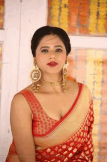 a woman in a red saree and gold earrings is standing in front of a wall .