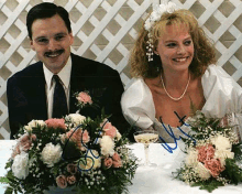 a bride and groom are sitting at a table with flowers and wine glasses .