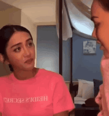 two women are talking to each other in a living room . one of the women is wearing a pink t-shirt .