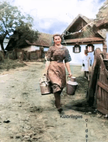 a woman carrying two buckets of milk is walking down a dirt road in front of a thatched roofed house