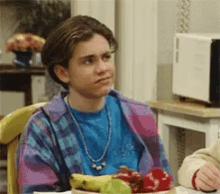 a young man is sitting at a table with a bowl of fruit and a microwave in the background .