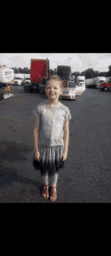 a little girl stands in a parking lot with trucks and a white truck that says ' morgan ' on the side