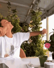 a woman is sitting at a table with her arms outstretched and wearing a white shirt that says nothing on it