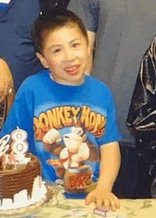 a young boy wearing a blue monkey mon shirt stands in front of a cake