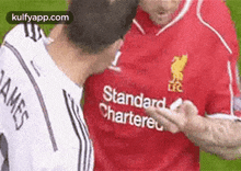 two men are standing next to each other on a soccer field . one of the men is wearing a red standard chartered jersey .