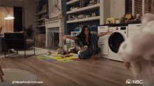 a woman is sitting on the floor with her arms outstretched in a living room with a nbc logo on the bottom right