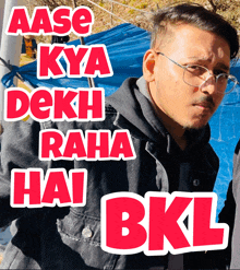 a man wearing glasses stands in front of a blue tarp with the words aase kya dekhi raha hai bkl