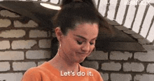 a woman in an orange shirt is standing in front of a brick wall and saying `` let 's do it . ''