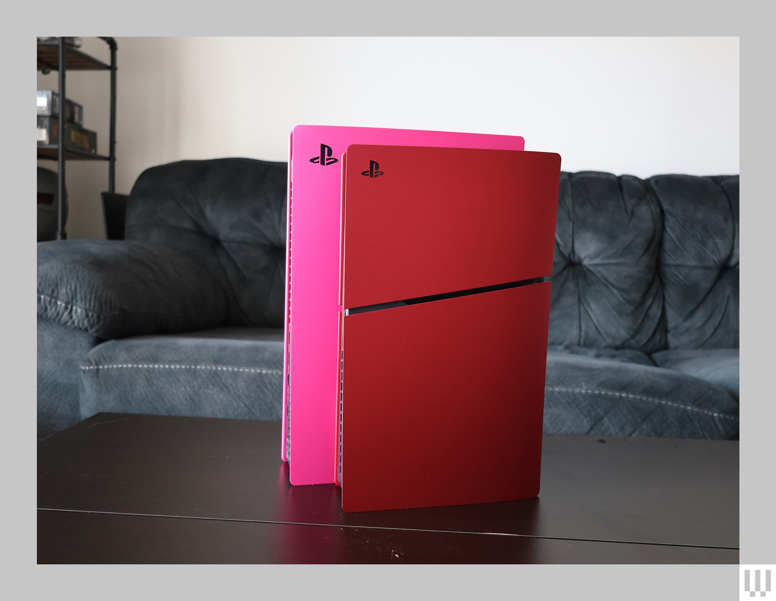 Two video game consoles with a pink one slightly behind a red one both sitting on a dark table with a couch in the...