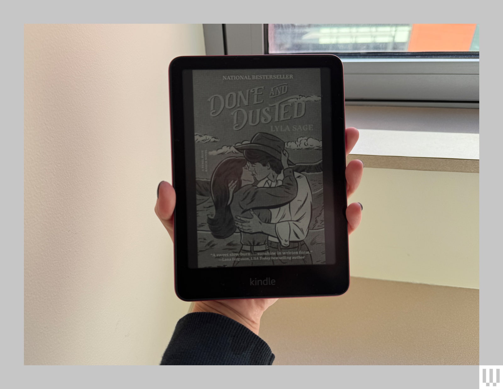 A hand holding up a Kindle Paperwhite a slim ereader with the screen showing the cover of an ebook