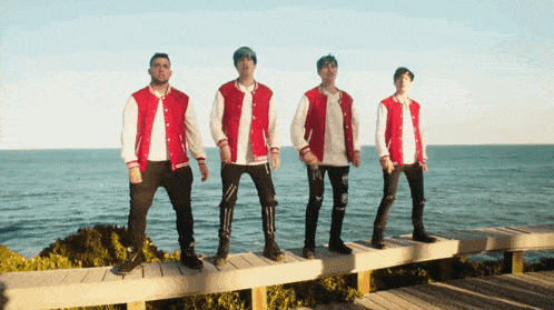 a group of young men wearing red and white varsity jackets are standing on a wooden dock overlooking the ocean