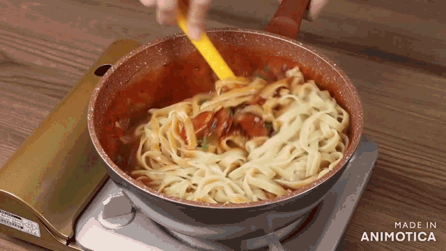 a pan of noodles is being stirred with a yellow spoon and the words made in animotica are visible in the corner