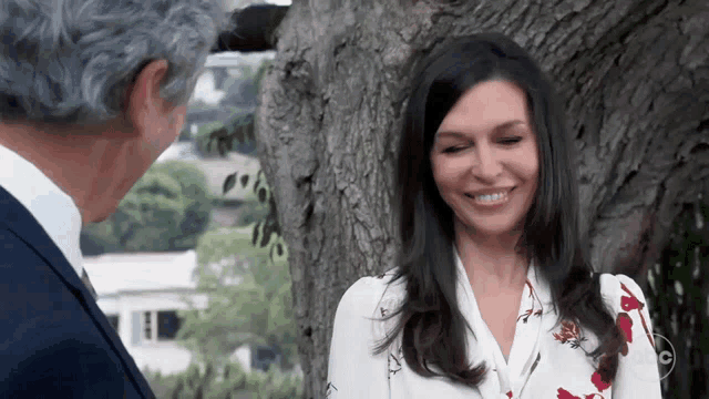 a woman in a floral shirt smiles while standing next to a man in a suit in front of a tree