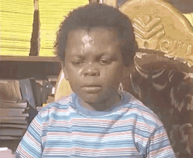 a young boy in a striped shirt is sitting in front of a bookshelf and making a funny face .