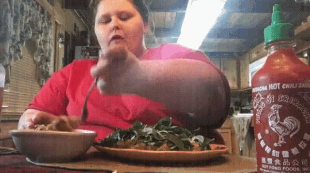 a woman in a red shirt is eating a salad with a bottle of sriracha hot chili sauce in the background .