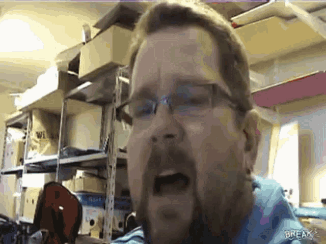 a man wearing glasses and a blue shirt is making a funny face in a room with boxes on shelves