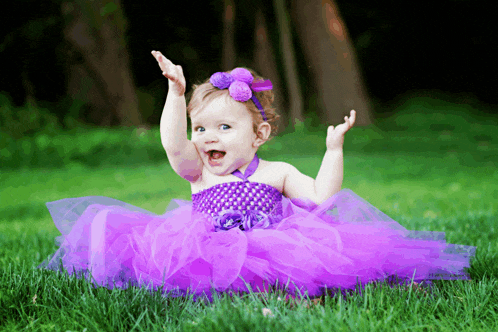 a little girl in a purple dress is sitting in the grass with her hands in the air