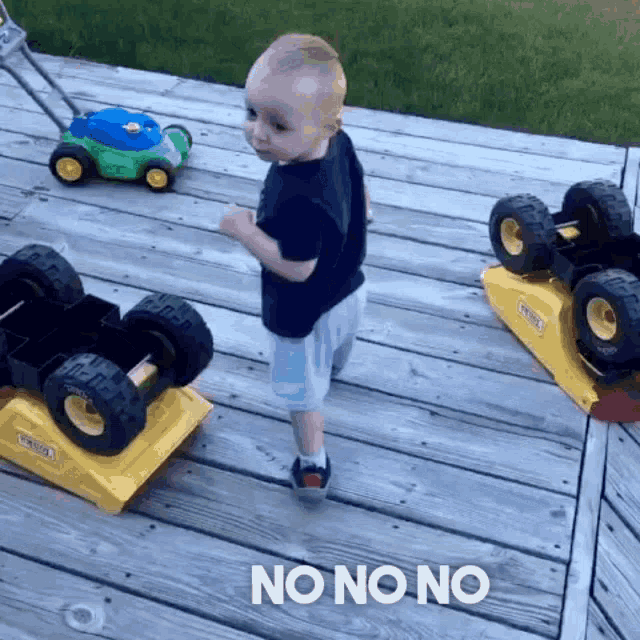 a little boy standing on a wooden deck next to a toy lawn mower and a toy truck that says no no no