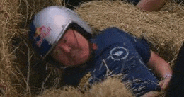 a man is laying in a pile of hay wearing a red bull helmet .