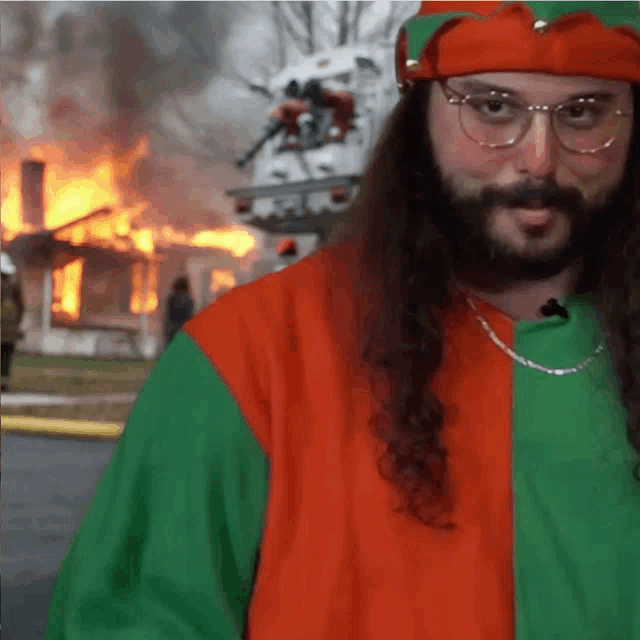 a man with long hair and glasses stands in front of a house that is on fire