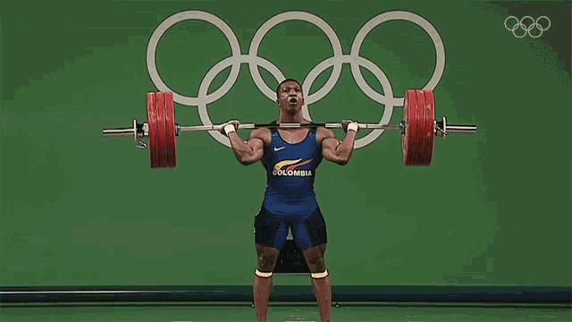 a man is squatting down while holding a barbell over his head with the olympic rings in the background .