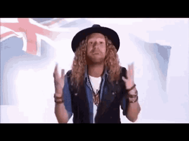 a man with long curly hair and a hat is giving the middle finger in front of an australian flag .