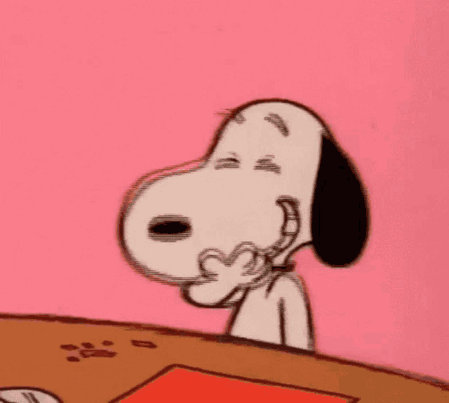 snoopy is sitting at a table and making a heart with his hands .