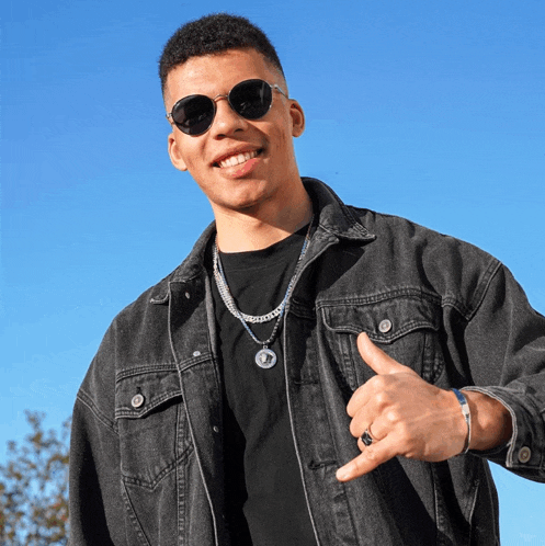 a young man wearing sunglasses and a denim jacket is giving a thumbs up