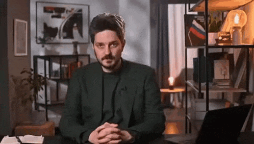 a man with a beard is sitting at a desk with his hands folded in front of him in a living room .