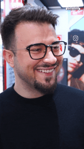 a man wearing glasses and a black shirt smiles in front of a picture of a man wearing sunglasses