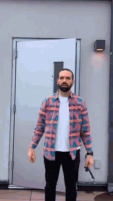 a man with a beard is standing in front of a door wearing a colorful shirt and holding a knife