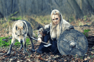 a woman is holding an axe and shield while a wolf watches .