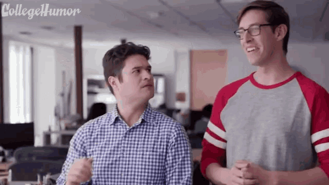 two men are standing next to each other in an office and one of them is wearing glasses and a baseball shirt .