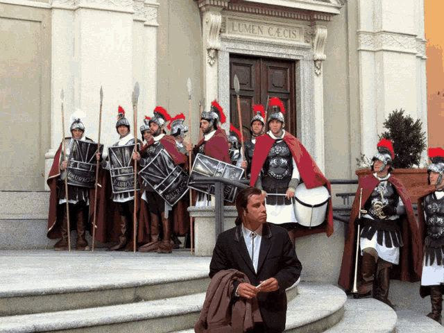 a man in a suit stands in front of a group of roman soldiers in front of a building that says lumen cecis