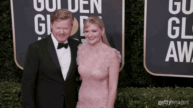 a man in a tuxedo and a woman in a pink dress pose on a red carpet in front of a golden globe sign