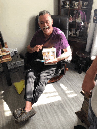 a man in a purple shirt sits in a chair holding a gift box