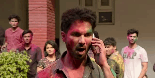 a man with holi paint on his face is talking on a cell phone in front of a crowd of people .