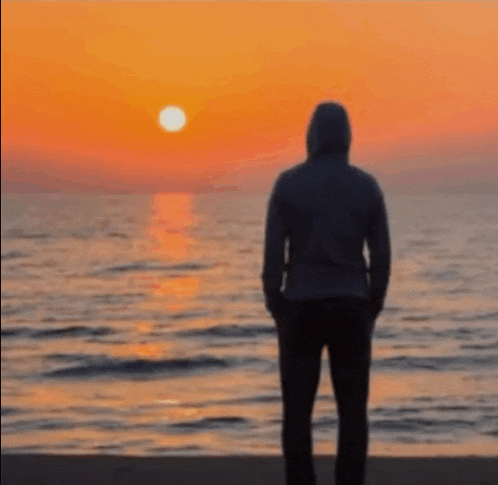 a man in a hoodie is standing on a beach at sunset looking at the ocean .