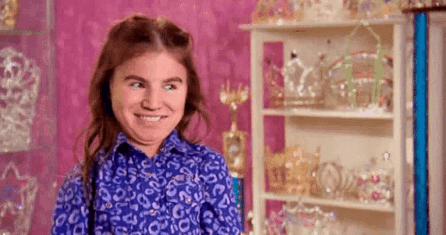 a young girl in a blue shirt is making a funny face in front of a shelf full of tiaras .