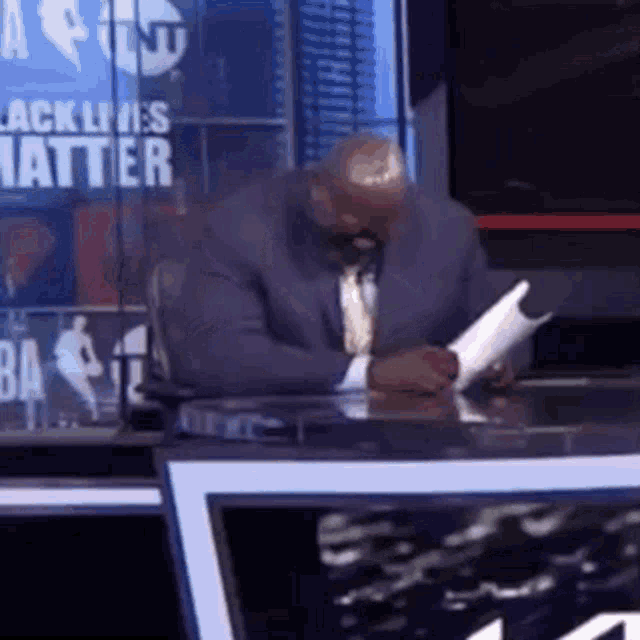 a man in a suit and tie is sitting at a desk in front of a sign that says black lives matter .