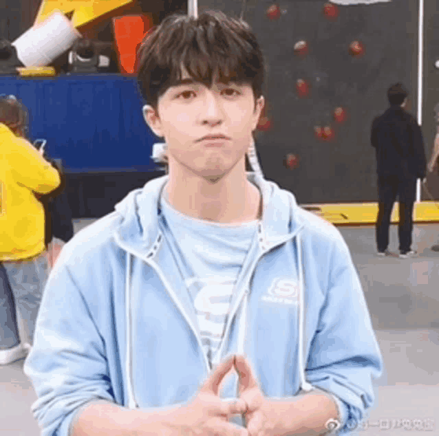 a young man in a blue sweatshirt is making a funny face while standing in front of a climbing wall .