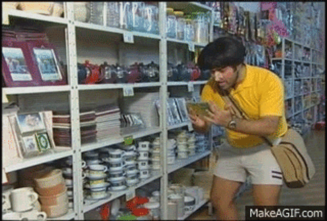 a man in a yellow shirt and shorts is standing in front of a shelf full of plates and cups .