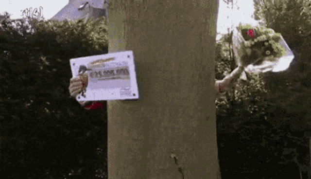 a person holding a bouquet of flowers behind a tree holding a sign that says ' loterij ' on it