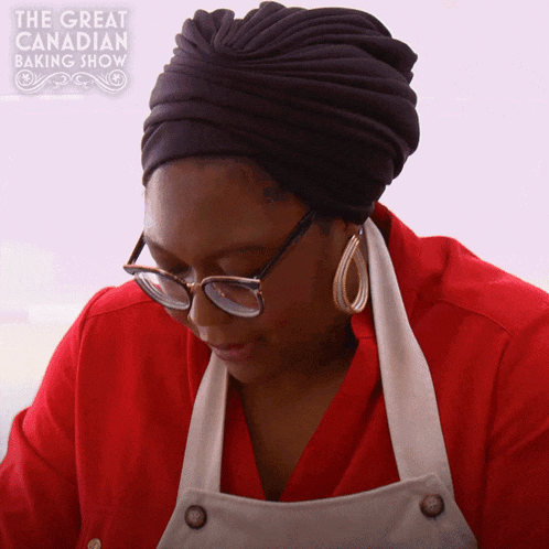 a woman wearing glasses and a turban looks down at something in front of a sign that says the great canadian baking show