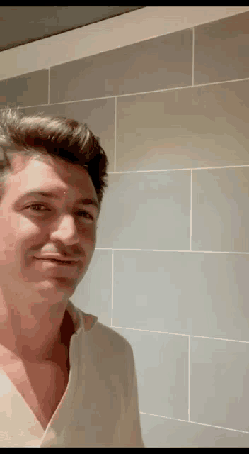 a man in a white shirt is standing in front of a tiled wall and smiling