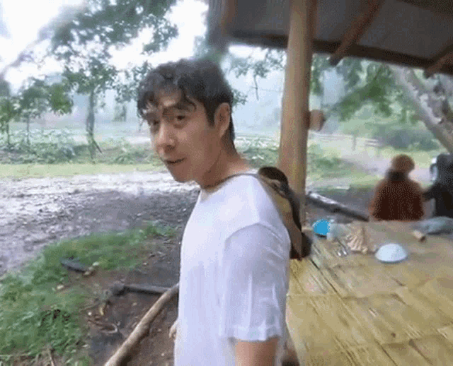 a man wearing a white shirt and a backpack is standing in front of a bamboo table in the rain .