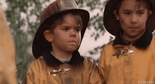 two young boys in firefighter uniforms are standing next to each other and one of them is wearing a fireman 's hat .