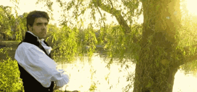 a man in a black vest and white shirt stands in front of a tree near a body of water