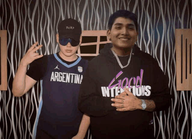 a man wearing an argentina jersey stands next to another man wearing a good intentions hoodie